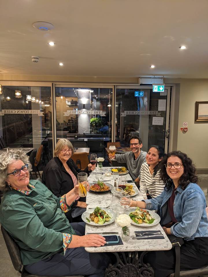 Five smiling travellers raise glasses over dinner inside a cozy modern restaurant.