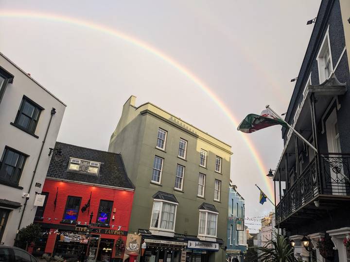 Rainbow arcs above colour-washed buildings and a Welsh flag along a charming town street at dusk.
