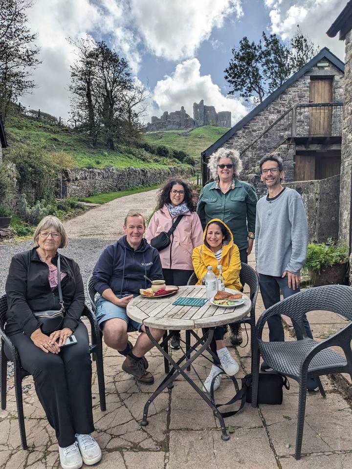 Travellers in casual outdoor gear relax around a café table with soup and pastries in a rural stone-walled setting.