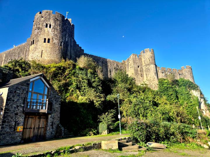 Sunlit stone castle rises above lush greenery and a converted cottage under a cloudless blue sky.