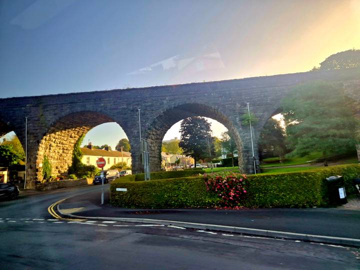 Stone railway viaduct with multiple arches spans a village road and manicured hedges in soft evening light.