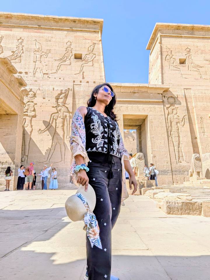 A woman wearing sunglasses poses in front of a sandstone Egyptian temple wall covered with large ancient relief carvings while other visitors mill about in the background.