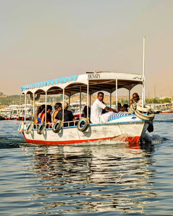 A small covered motorboat full of tourists cruises along a wide river with desert hills in the hazy distance.