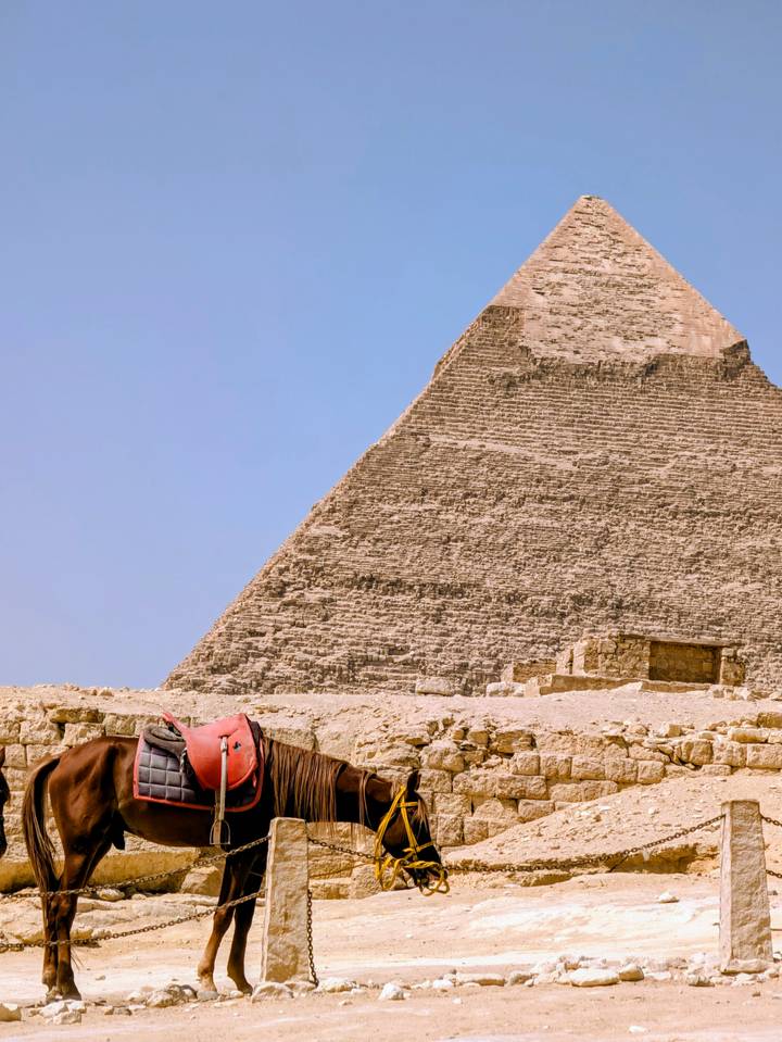 A massive stone pyramid rises from the desert with a resting camel and small stone hut in the sandy foreground under a clear blue sky.
