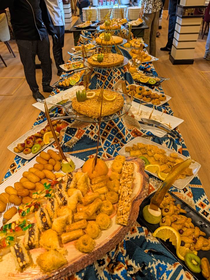 A lavish spread of Egyptian sweets and pastries is arranged on a patterned tablecloth inside a dining room.