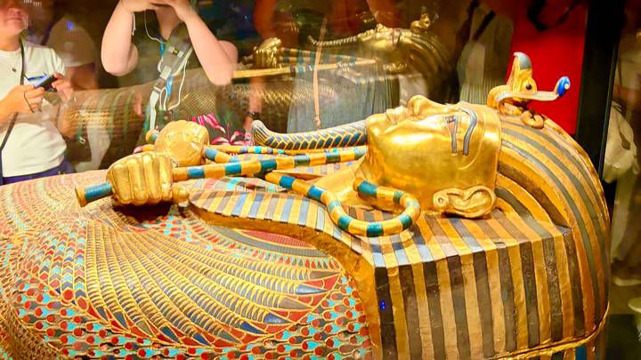 Gilded sarcophagus of an Egyptian pharaoh displayed behind glass with visitors looking on.