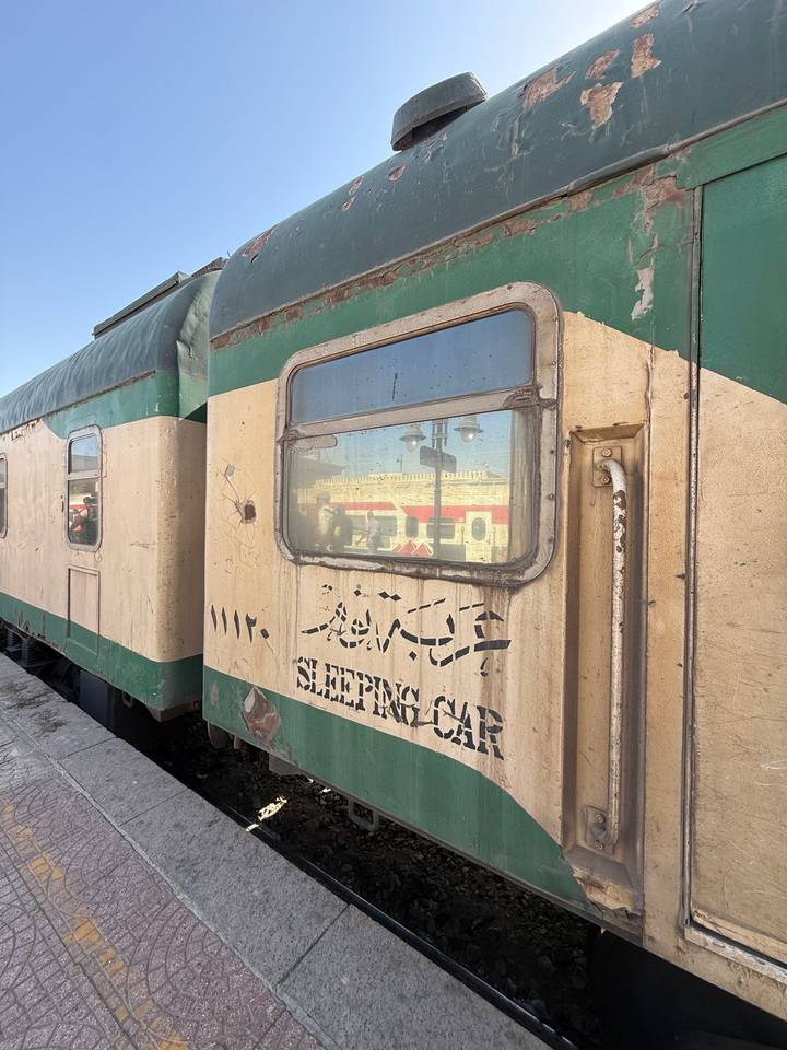 Weathered Egyptian sleeper train carriage window reflecting station platforms and locomotives.