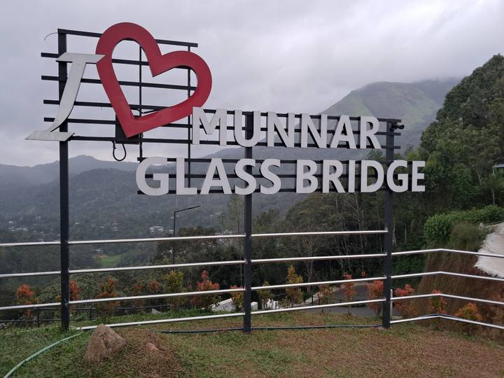 Large sign reading 'I ♥ Munnar Glass Bridge' set against misty green mountains
