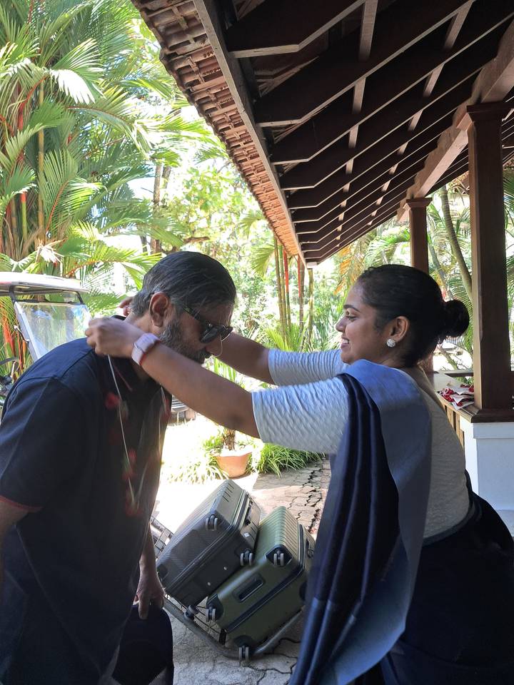 Woman placing a welcome garland on a guest at a tropical resort entrance surrounded by palms