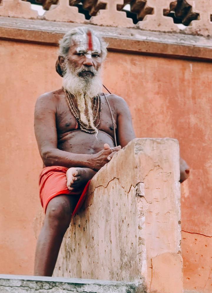 Close-up of a seated sadhu wearing prayer beads against an earthy terracotta wall.
