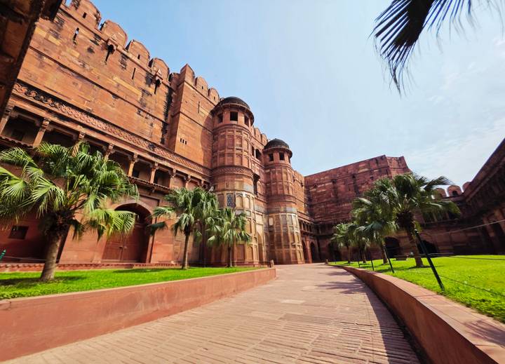 Wide courtyard of a red-sandstone fort flanked by palm trees and historic towers under bright sky.