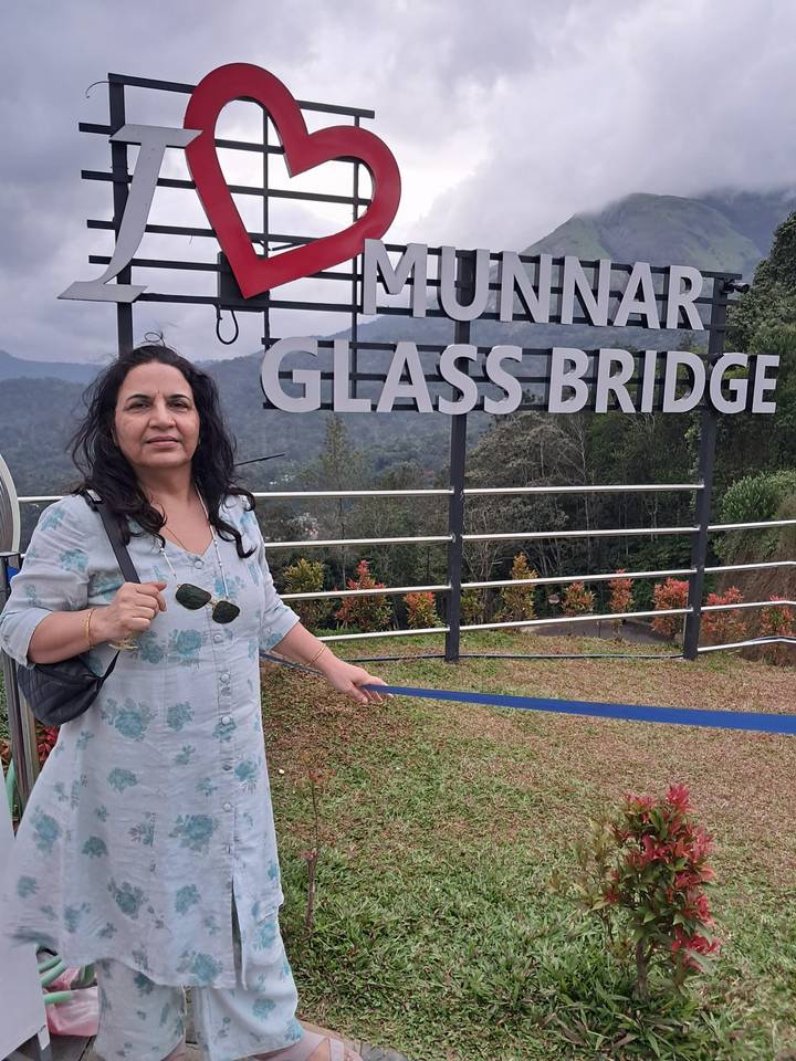 Woman holding sunglasses beside a sign reading "Munnar Glass Bridge" against misty hills.