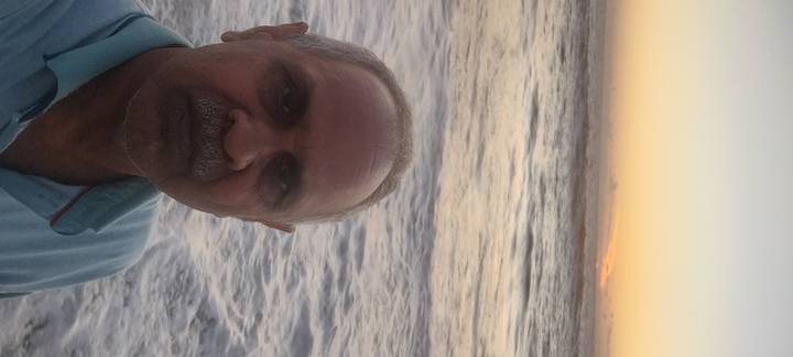 Self-portrait of an older man standing on a wave-washed beach at sunset with the sun dipping below the horizon.