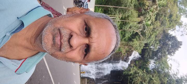 Selfie of a traveller with a roadside waterfall cascading behind him in a lush forested area.