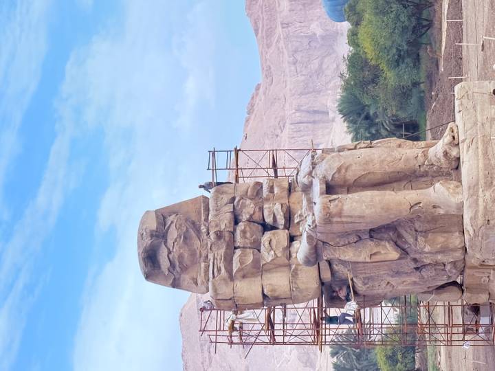 One of the Colossi of Memnon under restoration, with scaffolding and a worker against desert cliffs.