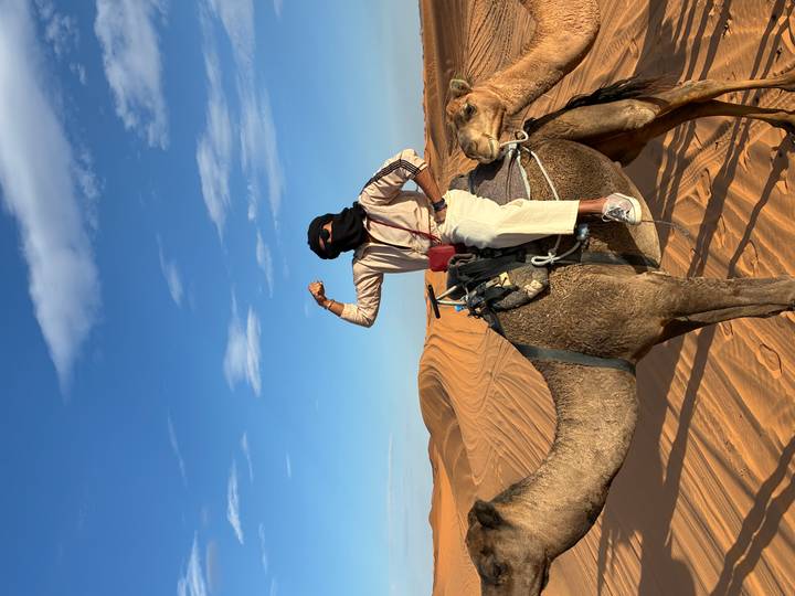Traveller confidently riding a camel across sweeping orange dunes under a blue sky.