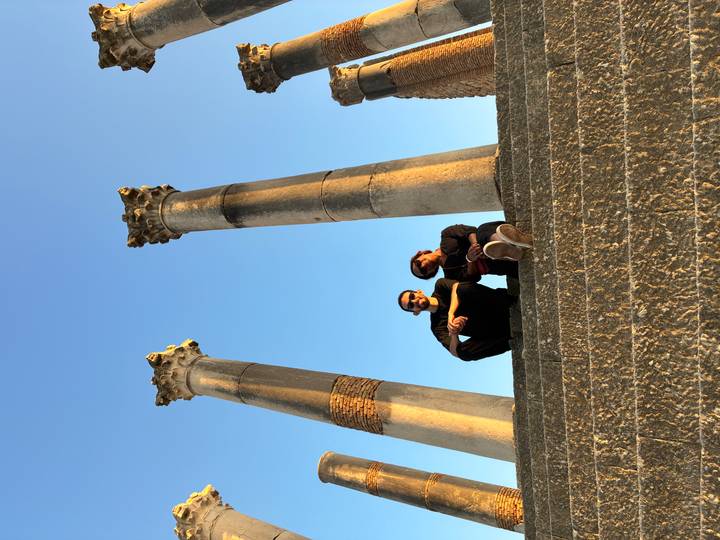 Two travellers sitting on stone steps beneath towering ancient Roman columns at Volubilis.