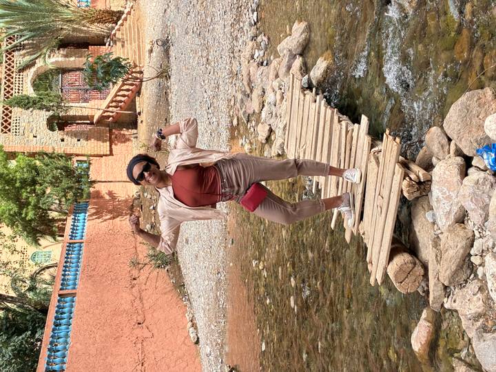 Traveller flexing on a makeshift wooden footbridge over a shallow mountain stream near an adobe village.