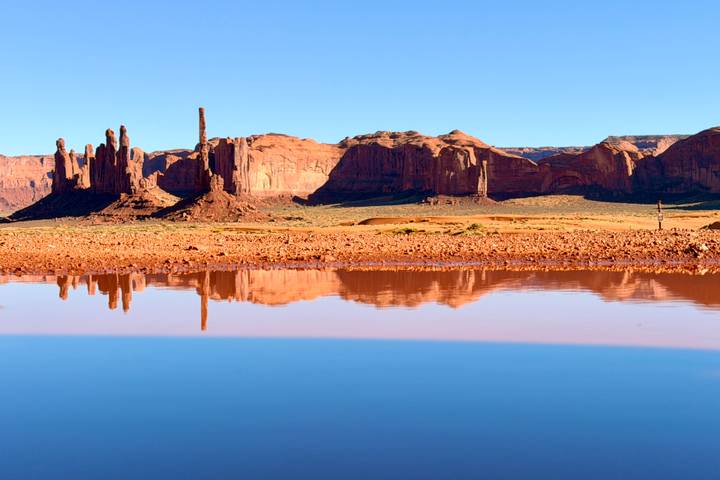 Red sandstone buttes of Monument Valley reflected in a still rain-fed pool under a clear blue sky.
