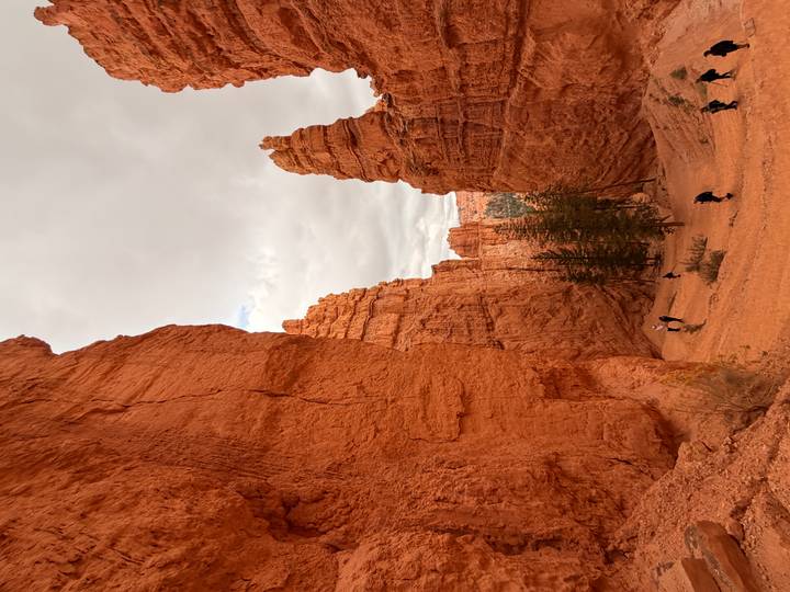 Visitors hike through towering orange hoodoos and narrow canyon walls under an overcast sky.