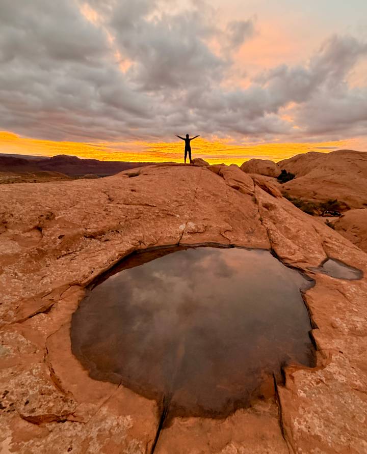 A lone traveller stands triumphant atop slickrock beside a rain-filled pothole reflecting the moody sunset sky.