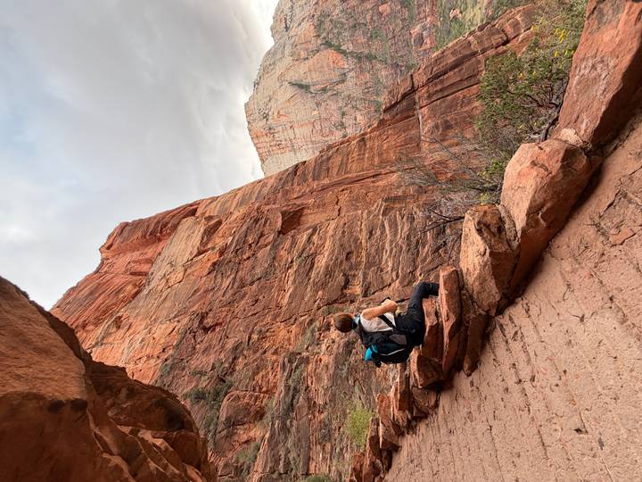A backpacker pauses on a narrow cliffside trail, gazing up at towering red canyon walls.