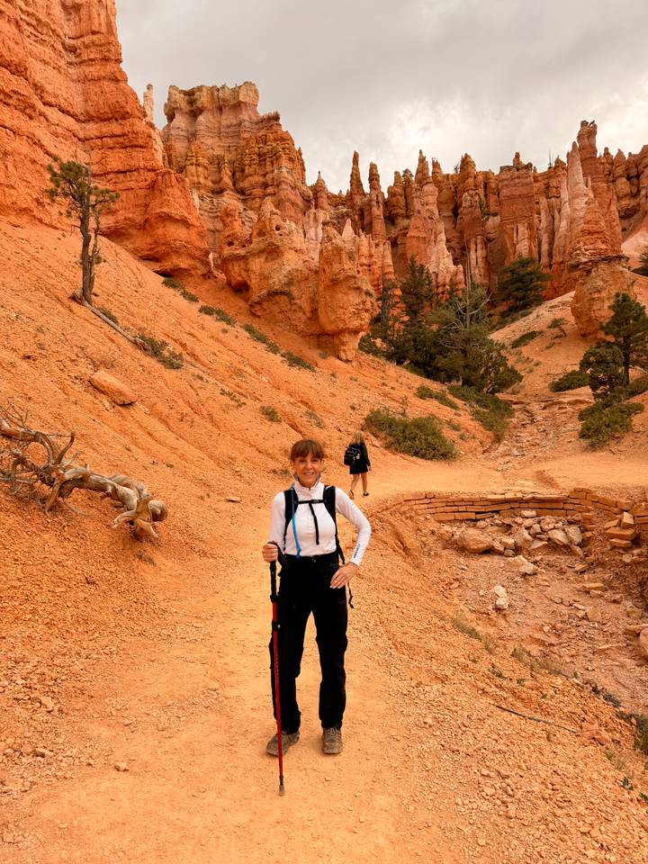 A smiling hiker poses with trekking poles along a dusty red canyon trail.