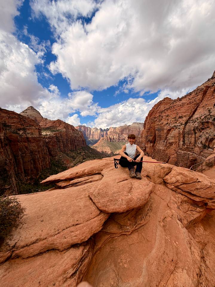A trekker rests on a cliff ledge overlooking dramatic canyon valleys beneath a patchwork sky.