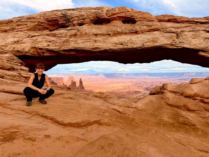 A traveller crouches beneath Mesa Arch framing vast desert mesas and spires beyond.