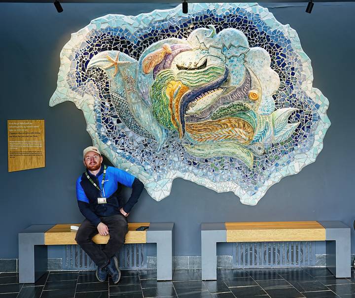Guide sitting beneath a colorful marine mosaic inside a visitor center