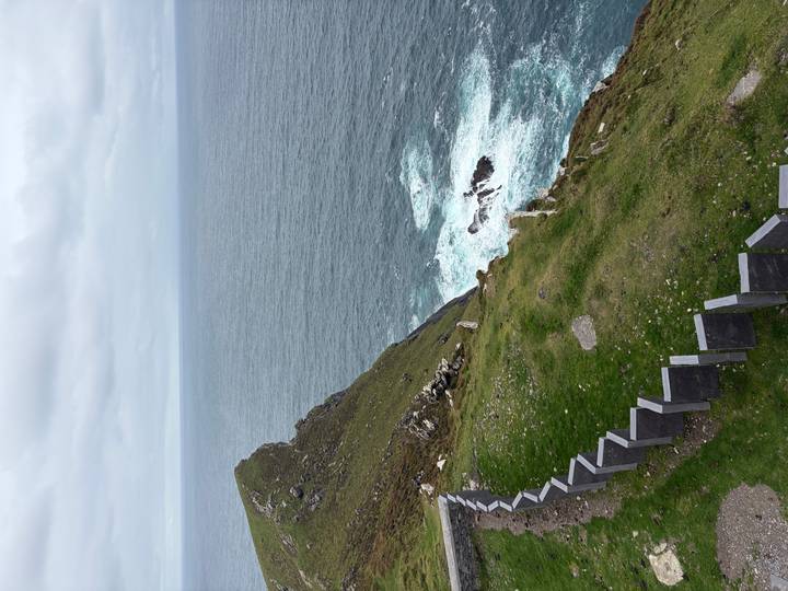 Steep grassy cliff dropping to the Atlantic with waves crashing below under gray skies