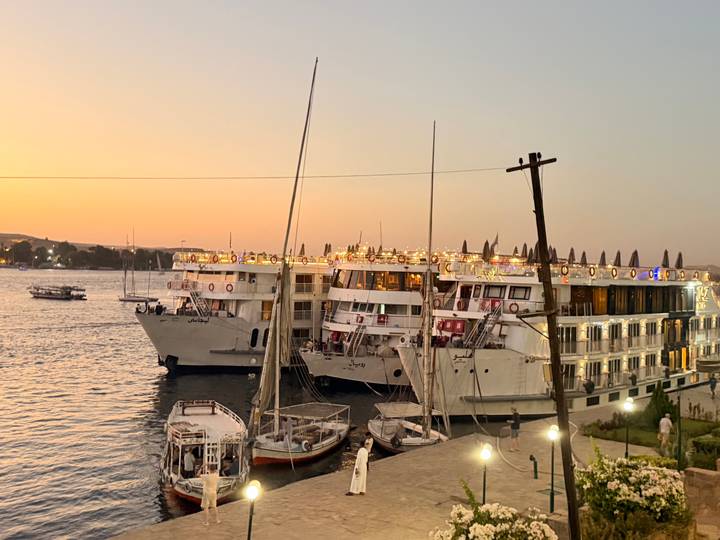 Nile cruise ships moored side-by-side glow with lights at sunset beside calm river water.