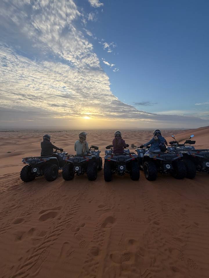 Five travellers on quad bikes look over sweeping Sahara dunes at sunset.