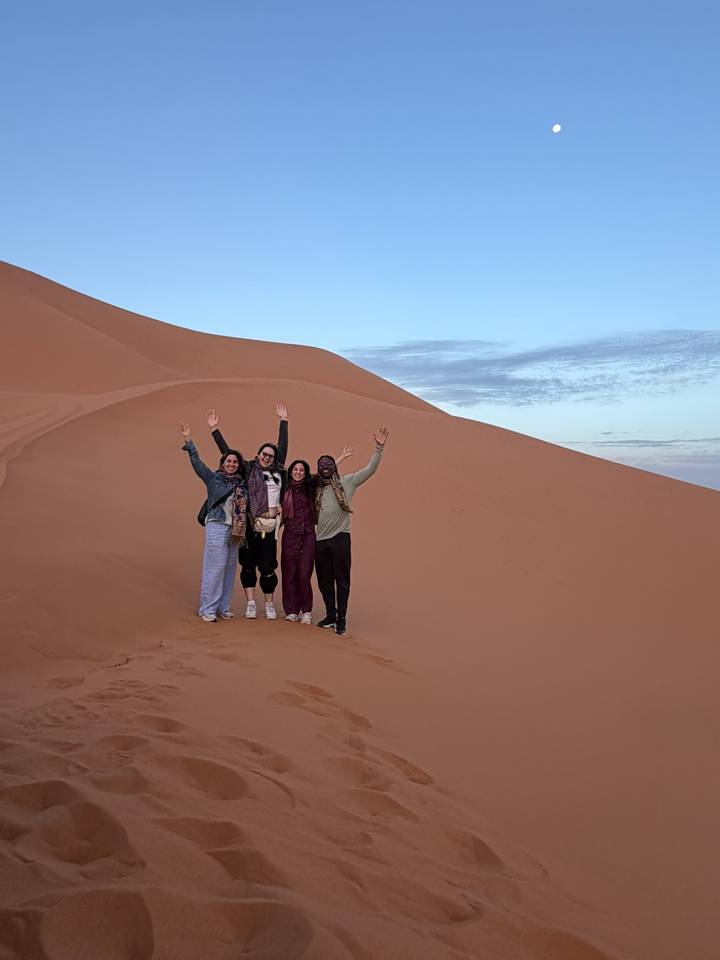 Happy group raises arms atop a tall orange dune in the Sahara at dusk.