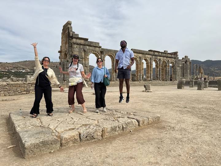 Travellers joyfully jump in front of ruined Roman arches on a dusty historic site.