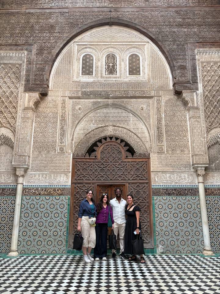 Group poses in front of intricately carved entrance of a medersa adorned with zellige tilework.