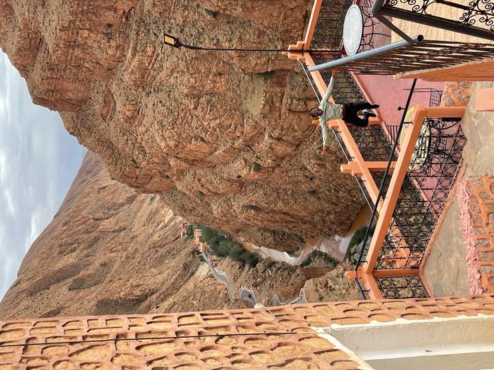 Traveler with arms outstretched on a terrace overlooking a winding mountain road and steep reddish canyon with a river below.