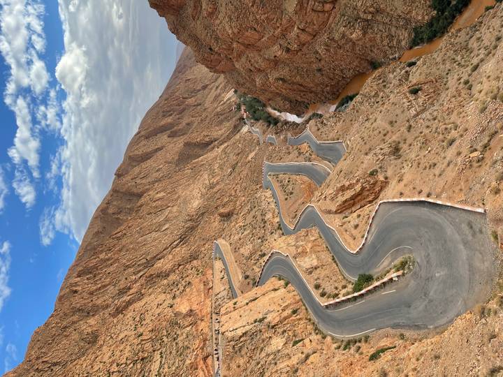 Serpentine mountain road carving through rugged reddish cliffs beneath a blue sky with scattered clouds.