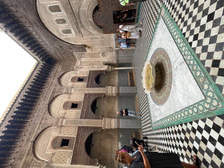 Mosaic-tiled courtyard with a marble fountain in a historic medersa, visitors standing along the walls.