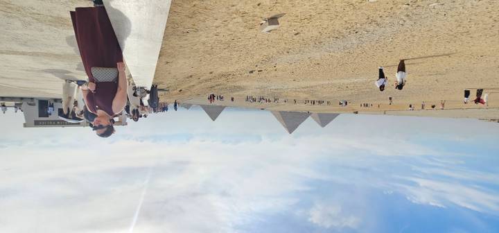 Wide desert panorama with distant pyramids and scattered tourists under hazy sky.
