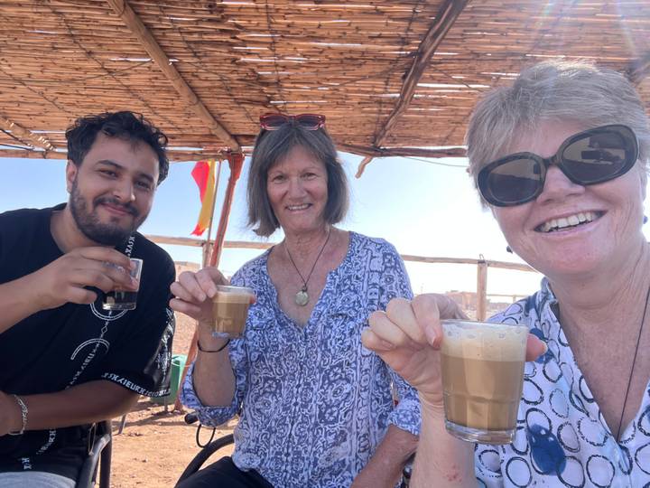 Three travellers smiling and toasting with frothy tea under a rustic shade structure in the desert.