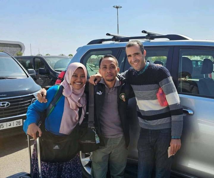 Three friends smiling for a photo in a parking lot beside parked vehicles.