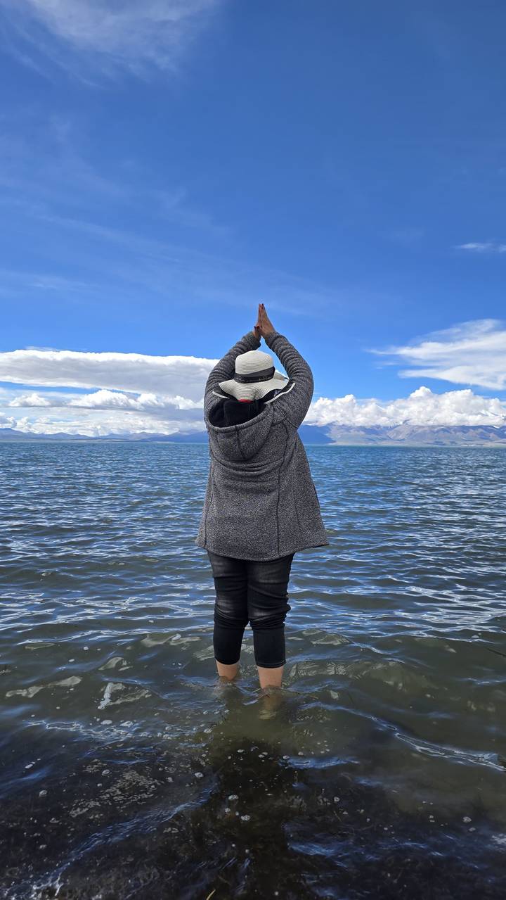 Person stands at a vast blue lake raising hands toward snow-capped Tibetan mountains.