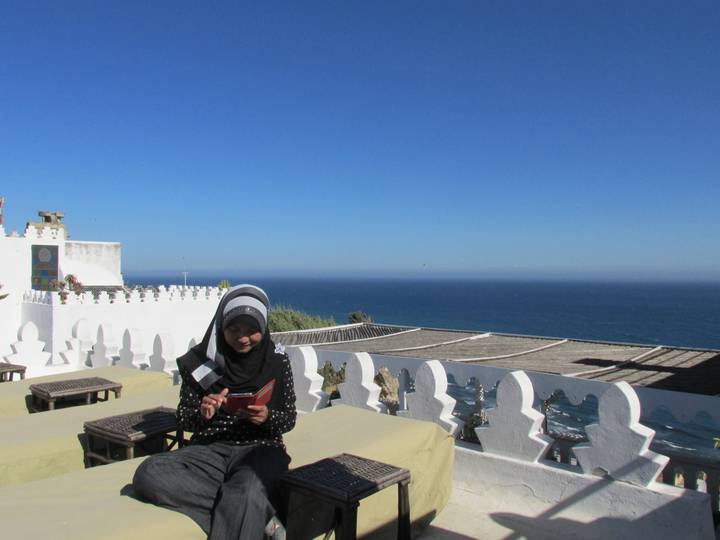 Young woman in hijab reads on a rooftop terrace overlooking a vast blue sea.