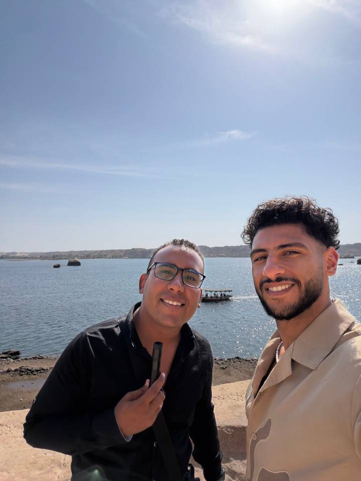 Selfie of two smiling travelers with a calm Nile backdrop and small motorboat.