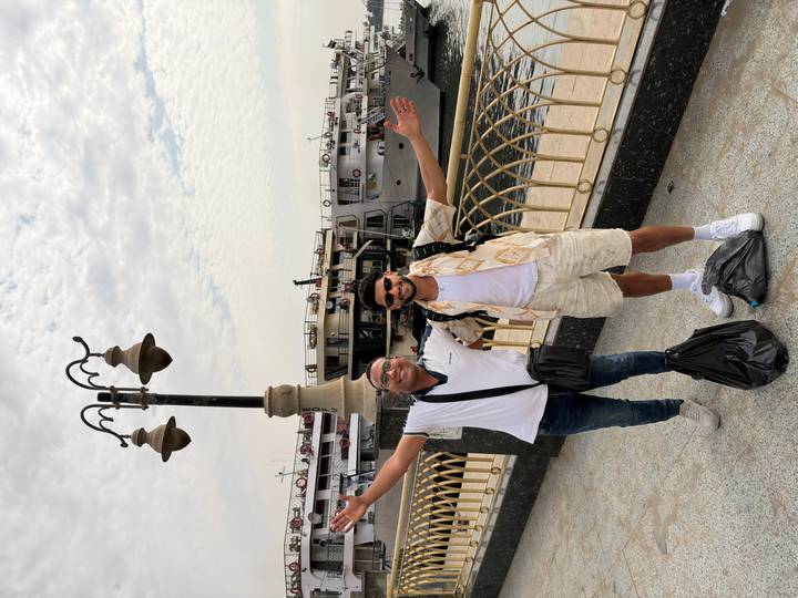 Two excited travelers spread their arms on a riverside promenade with cruise ships behind.