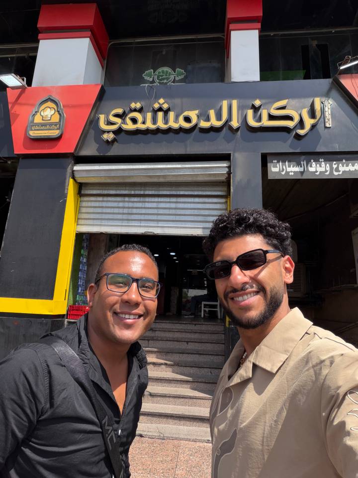Two friends smile in front of a storefront with Arabic signage and a closed metal shutter.