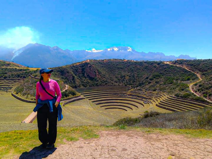 Woman stands before the concentric agricultural terraces of Moray with snow-capped Andes behind.
