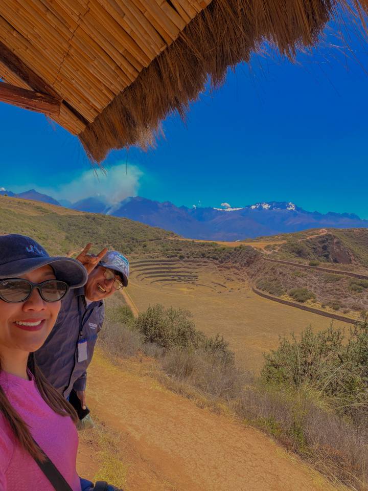 Selfie of two travelers smiling with the circular terraces of Moray and snow-peaked Andes in the background.