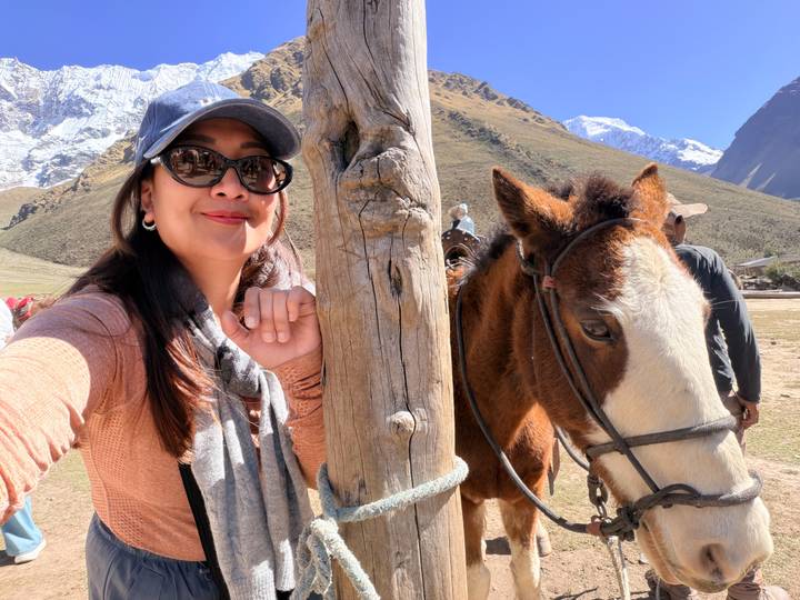 Smiling traveler beside a brown horse on a high-altitude mountain trail with snowy peaks behind.